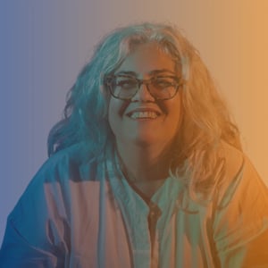 Mid 50s white woman with long grey hair and glasses smiling at camera