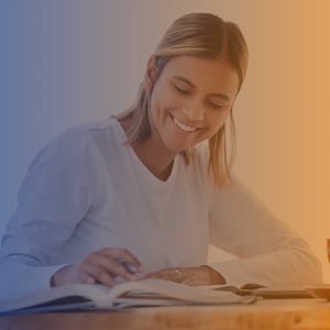Mid 30s white woman sat at table with open books, wearing a white top