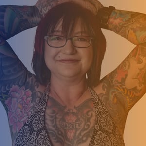 Mid 40s Asian woman wearing a patterned vest top with hands above her head showing full sleeve tattoos on both arms and across her chest